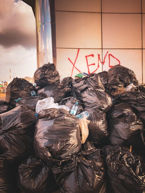 A large pile of black plastic rubbish bags, some torn open revealing plastic bottles and miscellaneous waste, stacked against an urban exterior wall with beige square tiles. Red graffiti spelling 'XEN